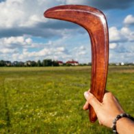 Close up view of a hand holding the Dark Brown Cedar boomerang against a green field.