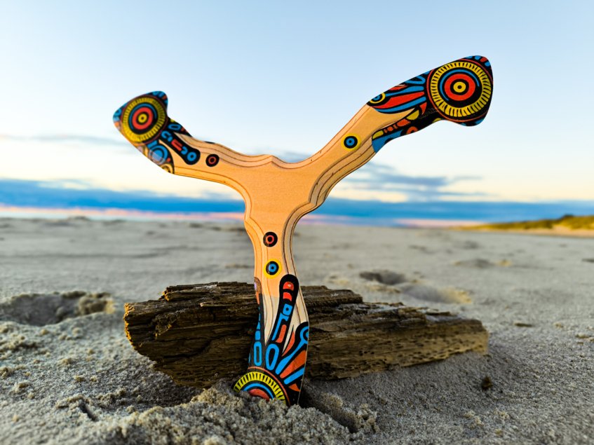 Colorful Seerblade wooden boomerang standing in beach sand next to driftwood.