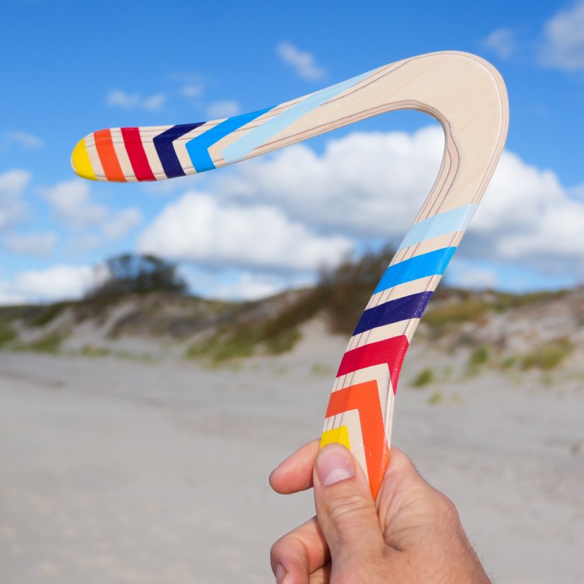 Skybreaker Real Wood Boomerang Sand The V-shaped Skybreaker wooden boomerang resting upright in beach sand under a cloudy sky.