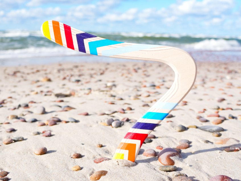 Skybreaker Traditional Wood Boomerang at the Beach The Skybreaker 2-wing returning wooden boomerang stuck playfully in the sand on a pebble beach.