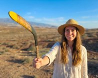 Smiling woman holding a traditional wooden boomerang in a sunny outdoor landscape.