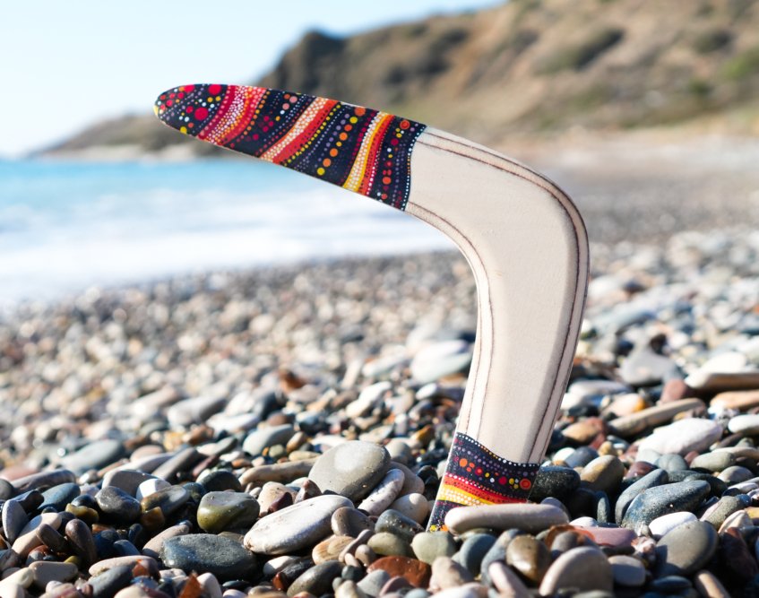 Golden Swallow wooden returning boomerang standing upright in beach pebbles with ocean background.