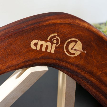 Custom Corporate Gift Boomerang with Engraved Company Logos Polished wooden boomerang with laser-engraved CMI and Compas Inc corporate logos displayed on a wooden stand