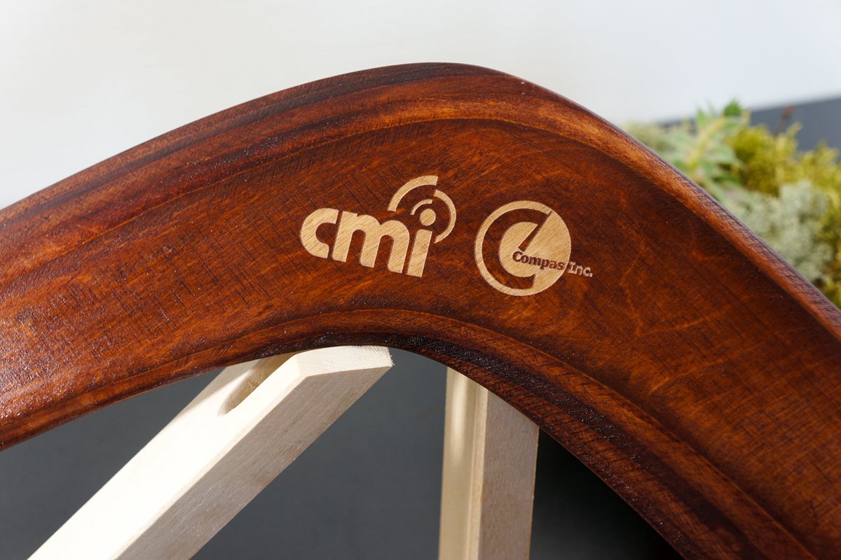 Polished wooden boomerang with laser-engraved CMI and Compas Inc corporate logos displayed on a wooden stand