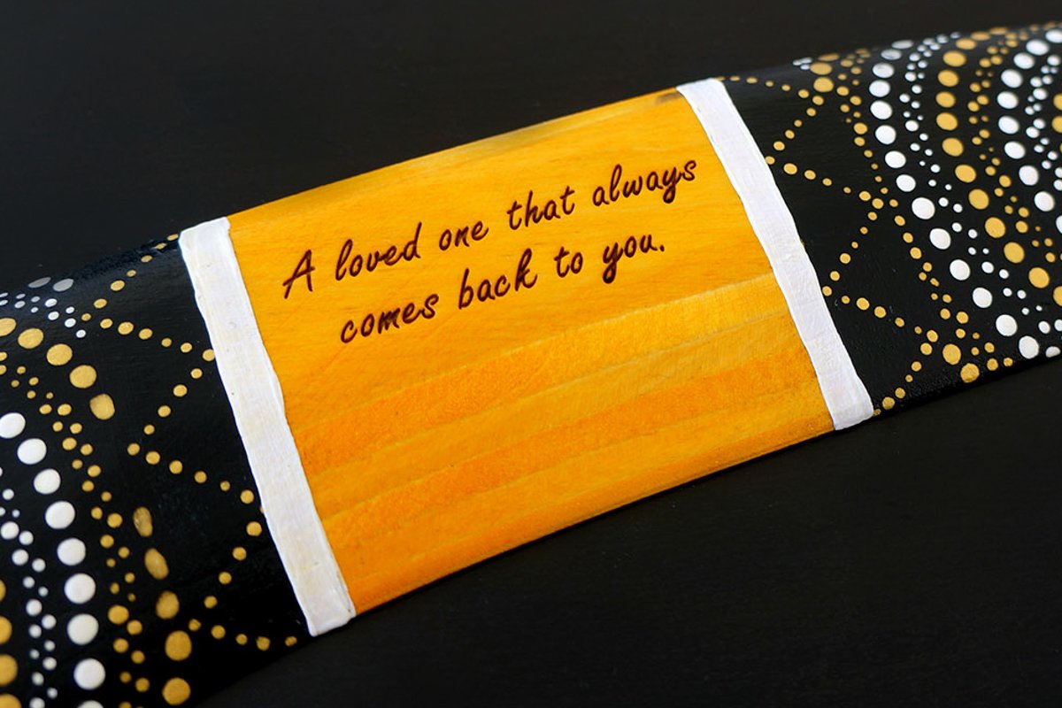 Handmade wooden boomerang with laser-engraved personal message on golden center panel surrounded by Aboriginal dot art in black white and gold